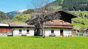 Ein traditionelles Chalet mit geneigtem Dach, Holzfassade und Steinfundament. Es wird von einem gepflegten Rasen und einer Bergkulisse umgeben.