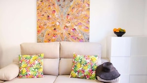 Un sofá color beige con dos almohadones estampados con motivos tropicales, un sombrero tejido oscuro y un cuenco de frutas sobre un mueble blanco.
