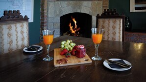A dining table with a fruit platter, two glasses of orange juice, and cutlery. A fireplace is visible in the background.
