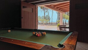A pool table with balls, a wooden bench, and a view of trees and a beach.