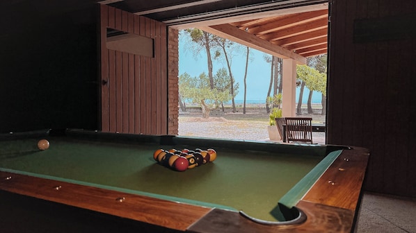 A pool table with balls, a wooden bench, and a view of trees and a beach.