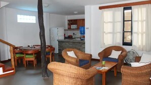 A cozy living room with wicker furniture, a dining area with wooden chairs, and a kitchen in the background.