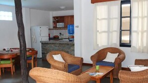 A living room with wicker furniture, a dining area with a wooden table, and a kitchen in the background.