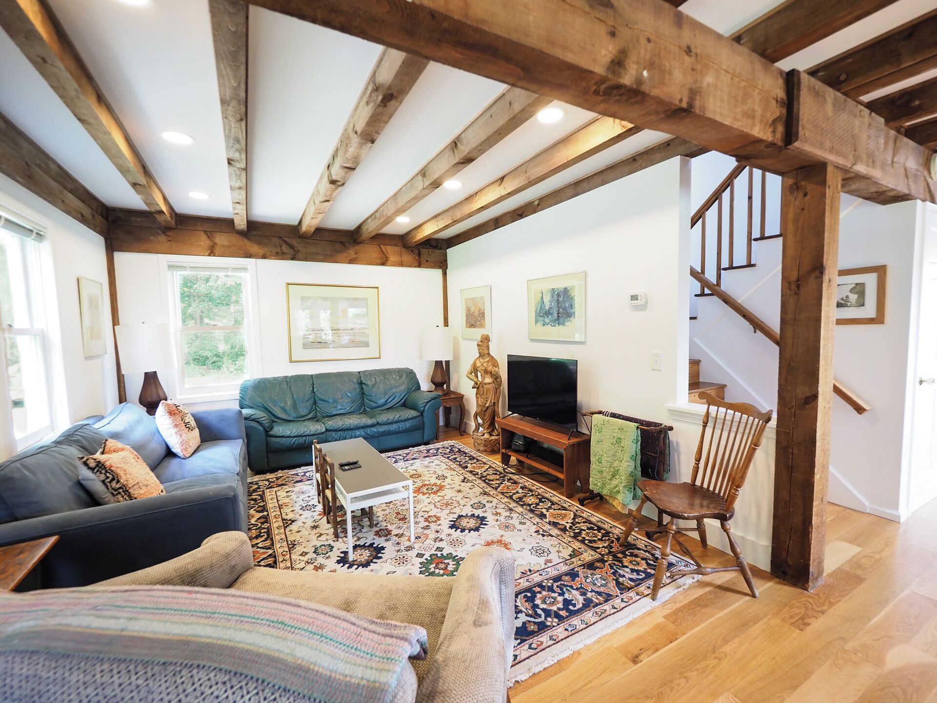 A cozy living room with a blue sofa, a patterned rug, wooden beams, and a staircase.