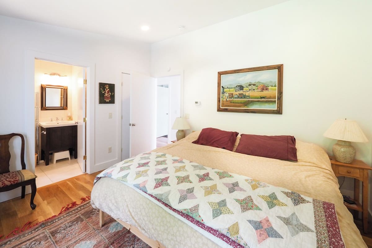 A bedroom with a large bed, a bedside table with a lamp, a painting on the wall, and a bathroom visible through an open door.