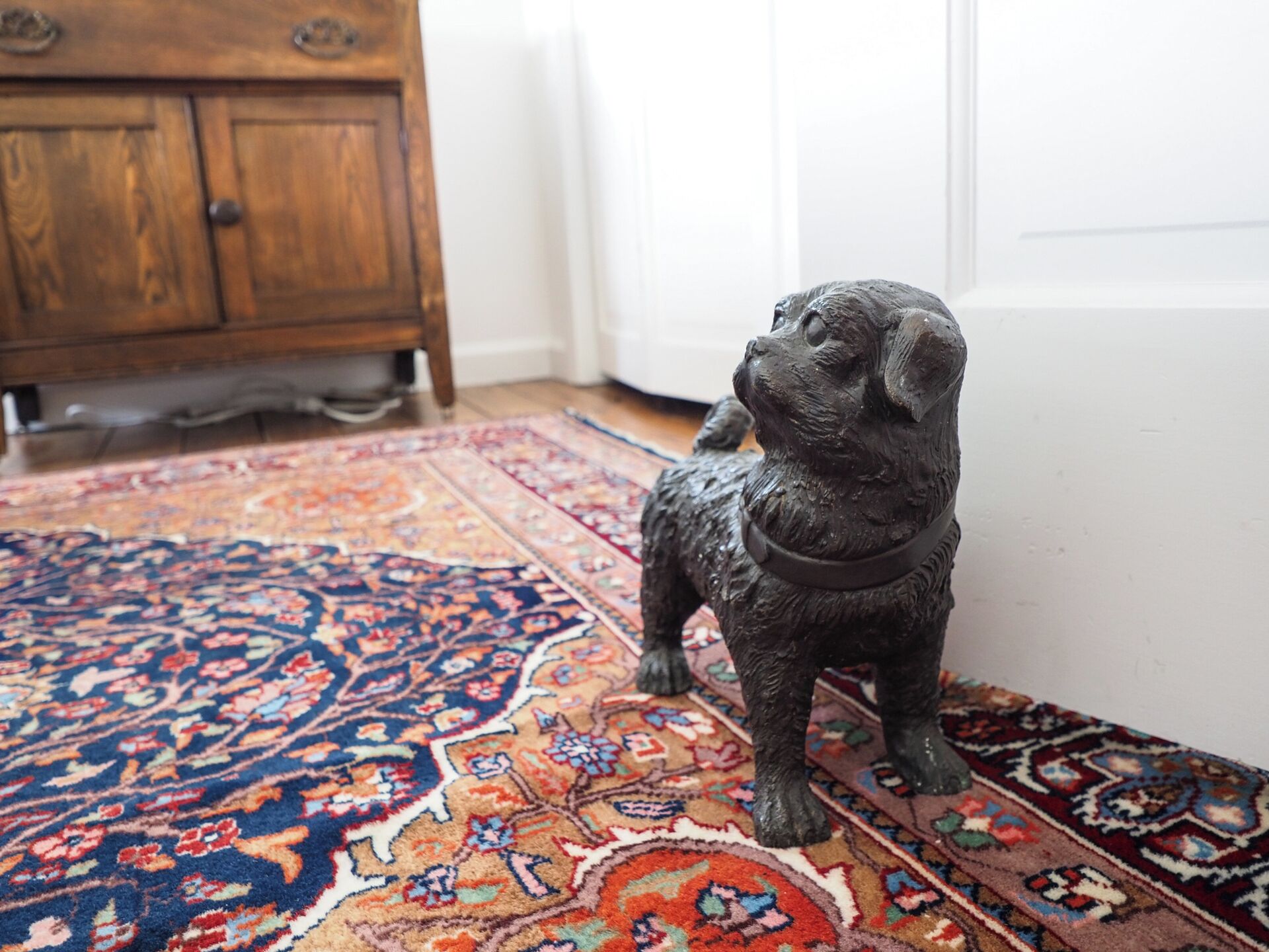 A small, dark-colored dog figurine on a patterned rug in a room with a wooden cabinet and a white door.