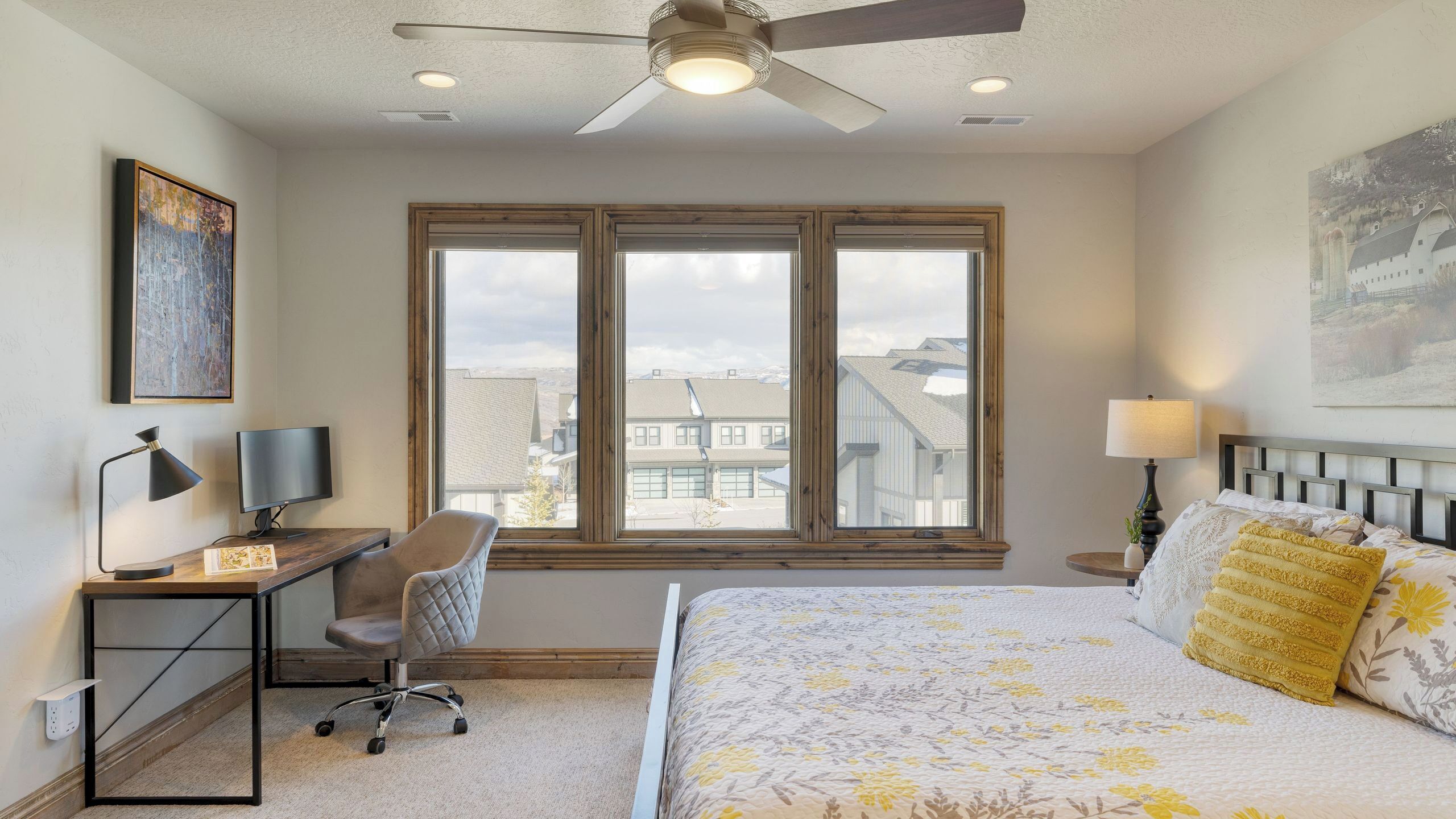 A bedroom with a bed, desk, chair, and a large window with a view of buildings.