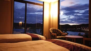 Habitación de hotel con dos camas, una silla, una mesita y vista a una masa de agua y edificios al fondo.
