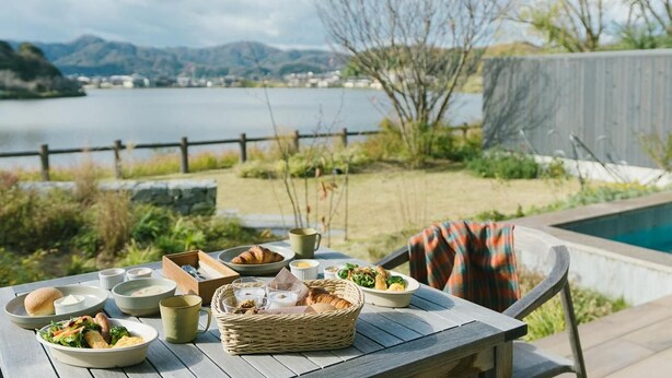 A dining table set outdoors with various dishes and a view of a lake and mountains. - Hotel & Lakehouse Soka (Kyotango)
