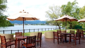 An outdoor seating area with tables and chairs, umbrellas, and a view of a lake and mountains. - Hotel & Lakehouse Soka (Kyotango)