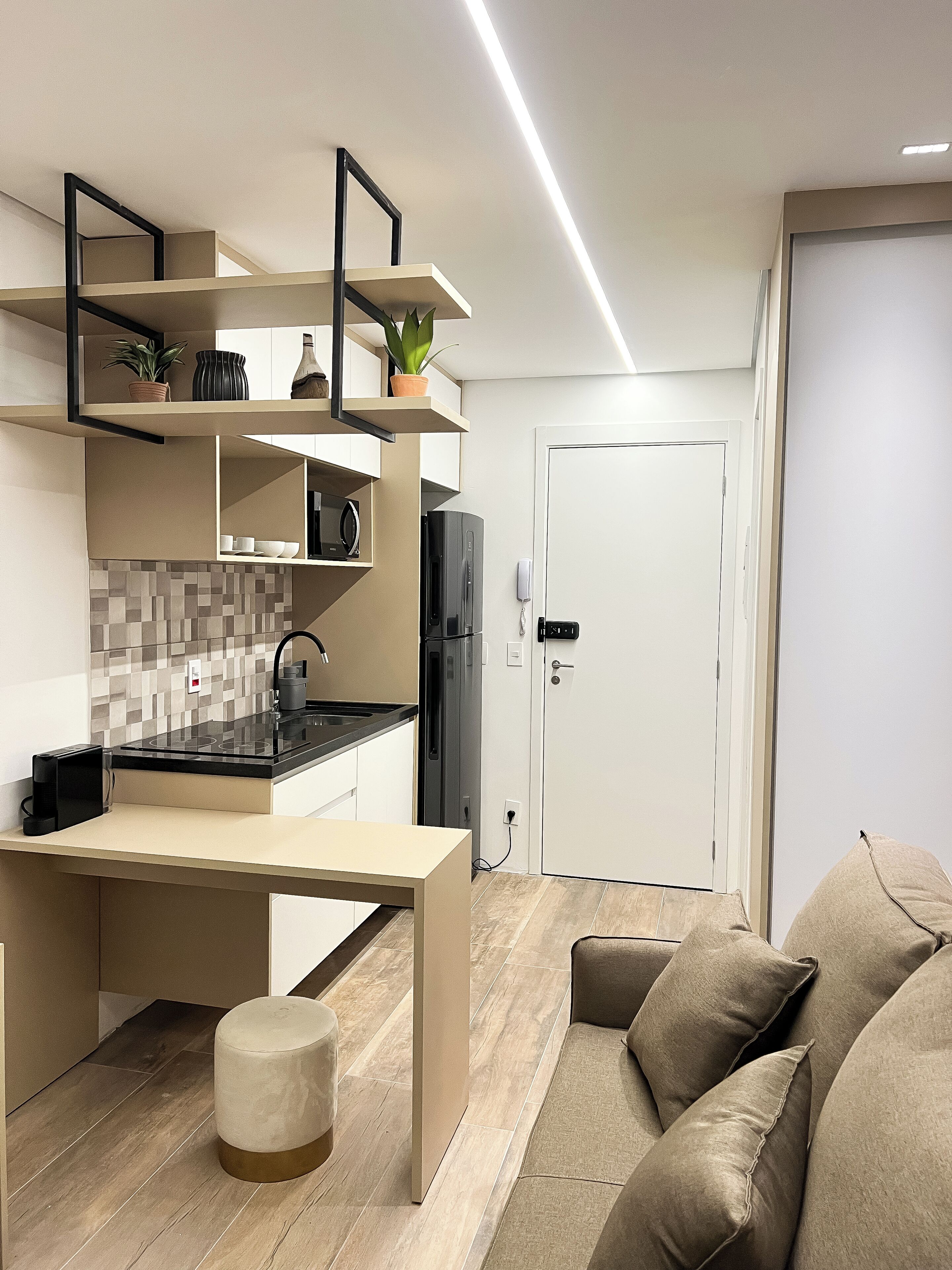 Una cocina moderna con encimera negra, armarios blancos y horno empotrado. Hay un taburete redondo pequeño y un sofá color beige en la sala de estar.