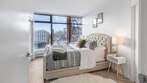 Ein modernes Schlafzimmer mit großem Fenster, einem gepolsterten Kopfteil, einem Nachttisch mit Lampe und Blick auf die Stadt.