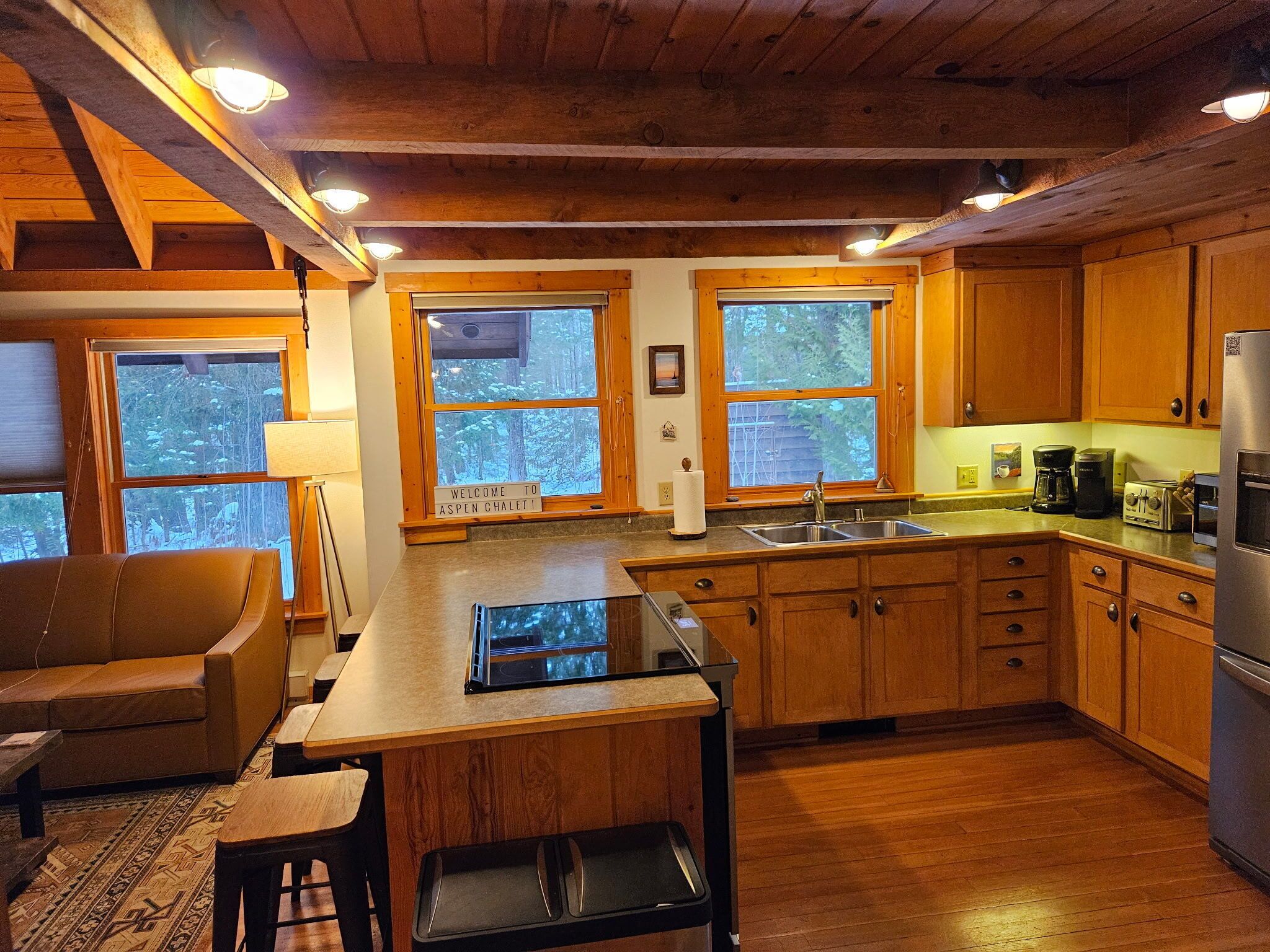 A cozy kitchen with wooden cabinets, a stainless steel sink, and a built-in stove. There are large windows with a view of trees outside.