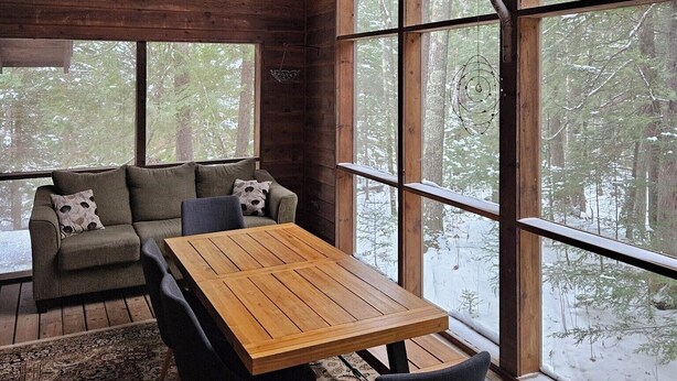 A cozy cabin interior with a wooden ceiling, large windows, a sofa, and a dining table.