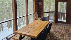 A wooden cabin interior with a large dining table, chairs, and a view of a snowy forest through large windows.