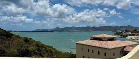 Une vue côtière avec un bâtiment doté d’un toit recouvert de tuiles et de nombreuses fenêtres, surplombant une mer calme et des montagnes lointaines sous un ciel partiellement nuageux.