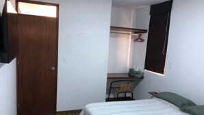 Um quarto pequeno e bem organizado, com uma cama, uma cadeira, uma escrivaninha e um guarda-roupa.