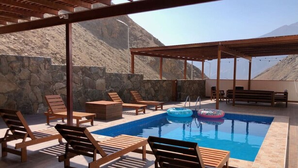 Área de piscina ao ar livre com espreguiçadeiras de madeira e um muro de pedra ao fundo.