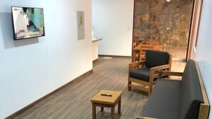 Quarto com mobiliário de madeira, incluindo um sofá e cadeiras, uma mesa pequena e uma televisão fixada na parede.