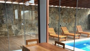 Mesa de jantar de madeira com cadeiras em um quarto com grandes portas de vidro que dão para uma parede de pedra e área da piscina.