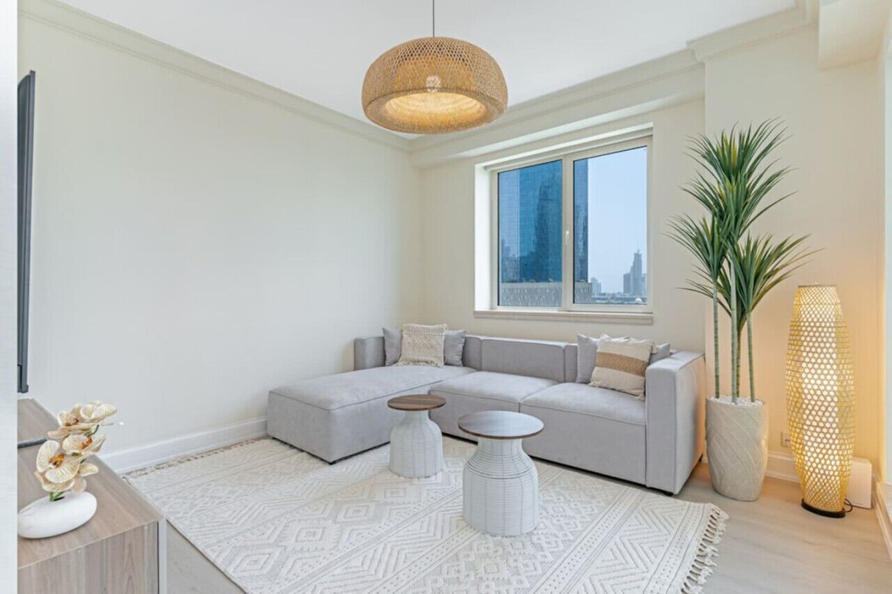 Ein modernes Wohnzimmer mit einem grauen Ecksofa, einem großen Fenster mit Blick auf die Stadt, einer gewobenen Pendelleuchte und einer hohen Topfpflanze.