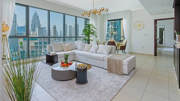 A modern living room with a large sectional sofa, a round coffee table with fruit, and a view of a city skyline through large windows. - Luxarious 2BR in Downtown Dubai with Infinity Pool and Burj Khalifa View! (Dubai)