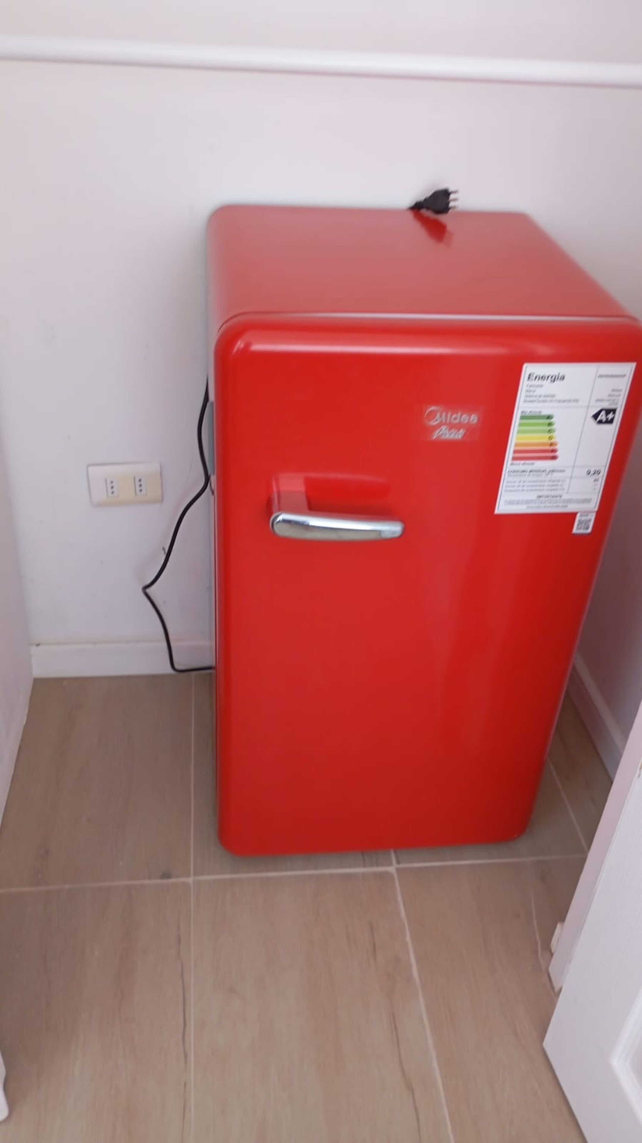 Un réfrigérateur rouge, doté d’une poignée, d’un câble d’alimentation et d’un autocollant indiquant les informations relatives à son efficacité énergétique.