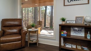 Ein Zimmer mit Ledersessel, kleinem Tisch mit Pflanze, Bücherregal mit Büchern und einem Willkommensschild, sowie einem großen Fenster mit Blick auf Bäume.