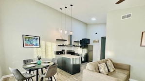 A modern kitchen with stainless steel appliances, a dining area with a round table and chairs, and a living area with a sofa and decorative pillows.