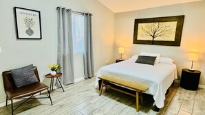 A bedroom with a bed, bedside lamps, a bench, a chair, a small table with flowers, and a framed picture on the wall.