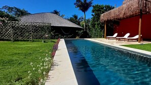 A swimming pool with lounge chairs, a thatched-roof structure, and a red building. - Trancoso. Alto do Segredo. 2 Suites.  5 minutes from the beach and 2 km from Quadrado (Trancoso)