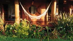 A hammock hanging between wooden pillars in a garden area with a view of a building with a circular window and a door. - Trancoso. Alto do Segredo. 2 Suites.  5 minutes from the beach and 2 km from Quadrado (Trancoso)