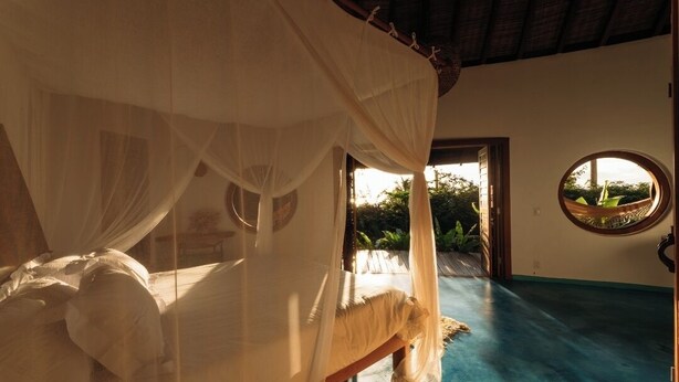 A room with a bed under a canopy, a round window, and a view of greenery outside. - Trancoso. Alto do Segredo. 2 Suites.  5 minutes from the beach and 2 km from Quadrado (Trancoso)