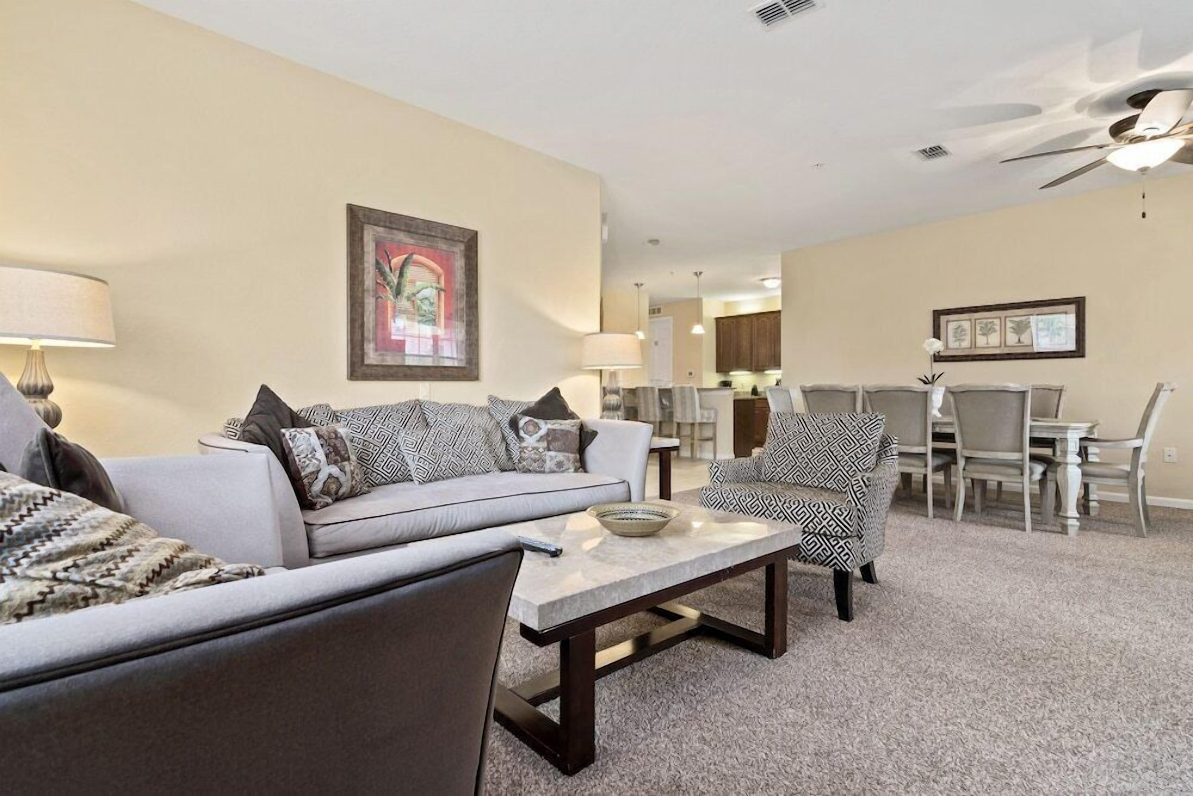 A living room with a sofa, armchair, and coffee table, a dining area with a table and chairs, and a kitchen in the background.