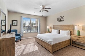 A hotel room with a bed, bedside tables, a desk, a chair, a window with curtains, a ceiling fan, and a wall decoration. - Orlando Resort Rentals Near Universal (Orlando)