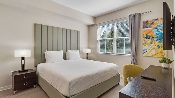 A modern hotel room with a bed, bedside tables, a desk, a chair, a TV, and a window with blinds. - Orlando Resort Rentals Near Universal (Orlando)