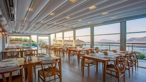 Une salle à manger spacieuse, dotée de grandes fenêtres offrant une vue sur une étendue d’eau et sur le paysage lointain. Les tables sont dressées avec des nappes blanches et des chaises en bois.