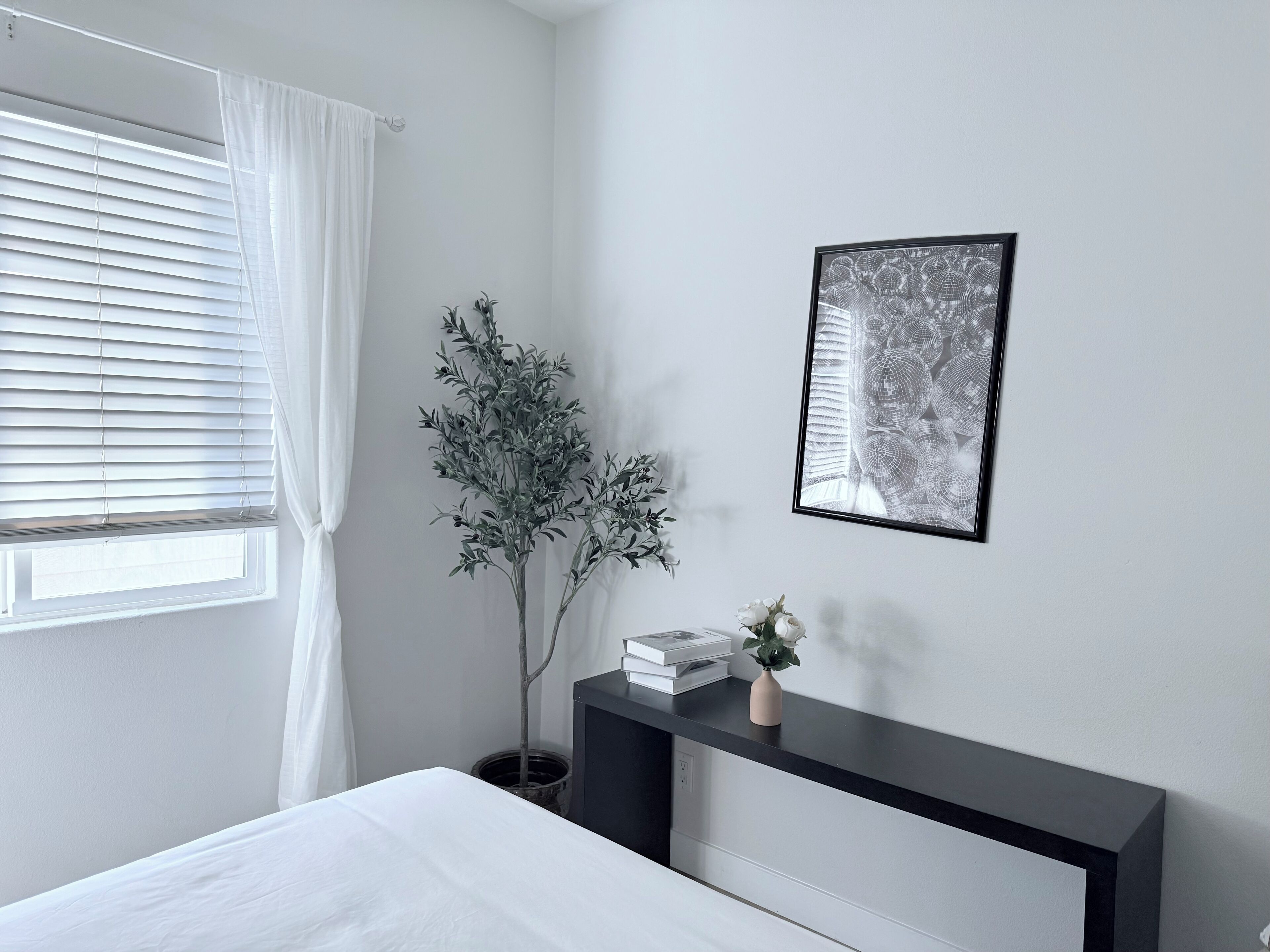 Un dormitorio con cama, una mesita de noche con un jarrón y libros, una planta en maceta, una ventana con persianas y un cuadro en la pared.