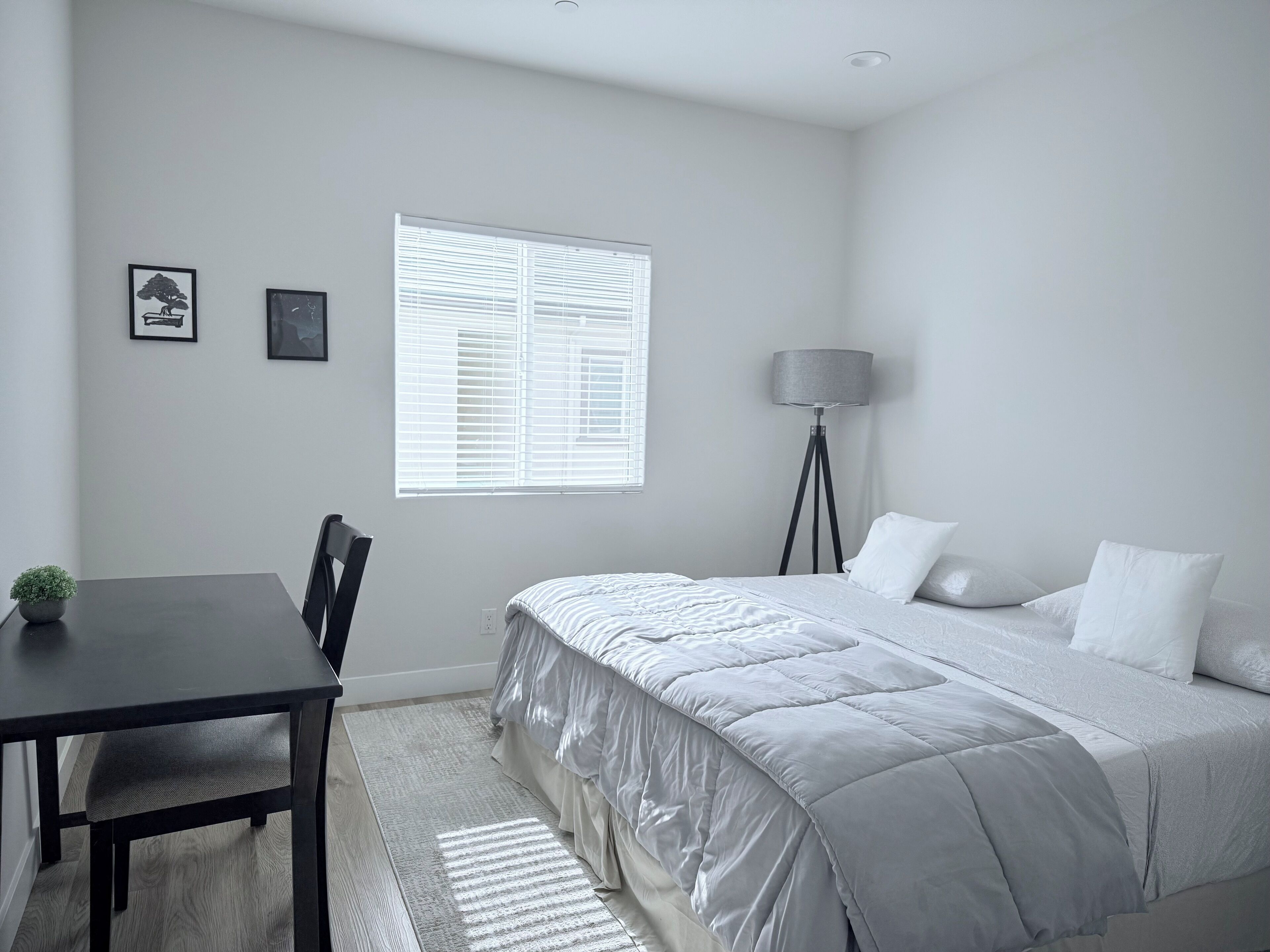 Une chambre à coucher comprenant un lit, un bureau, une chaise, une lampe sur pied et deux tableaux encadrés accrochés au mur.