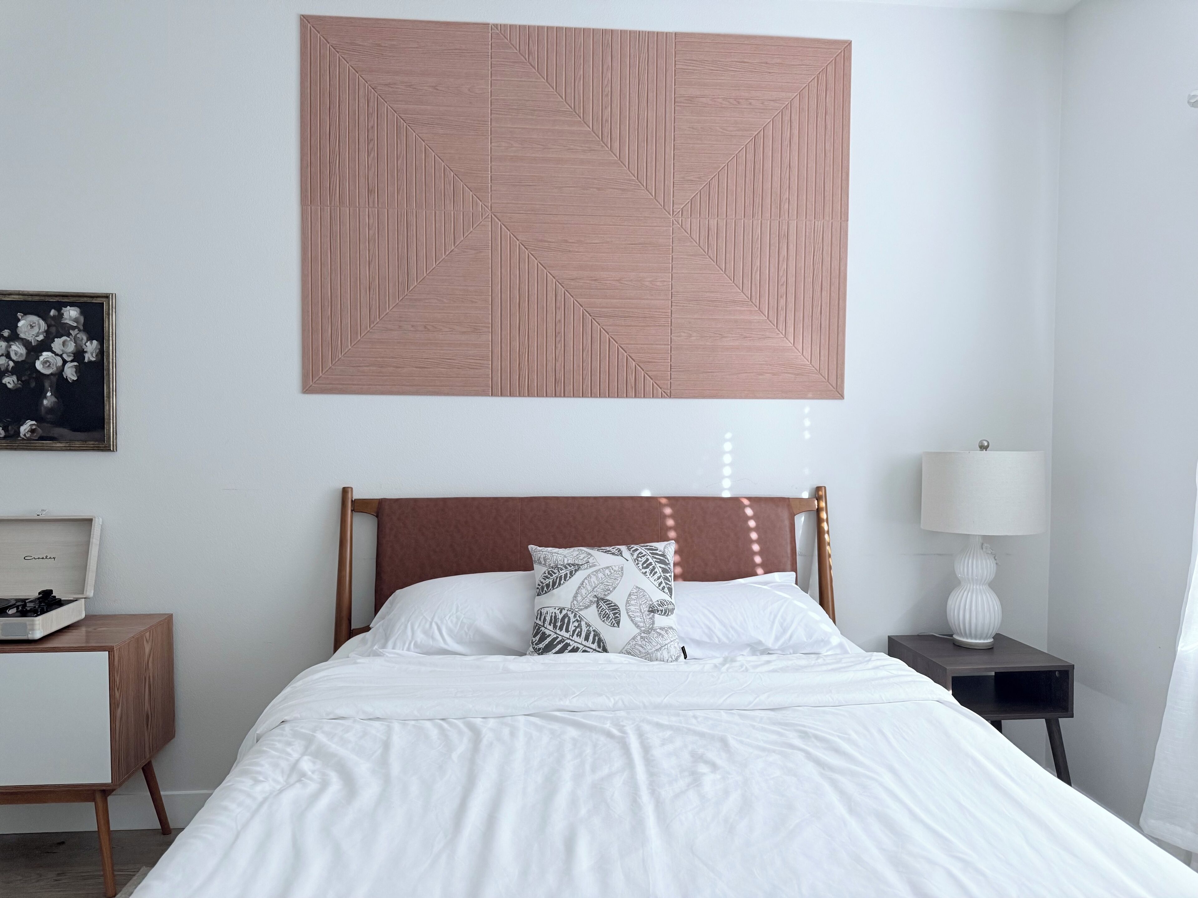 A neatly arranged bedroom with a large bed, a bedside table, a nightstand, a wall-mounted artwork, and a lamp.