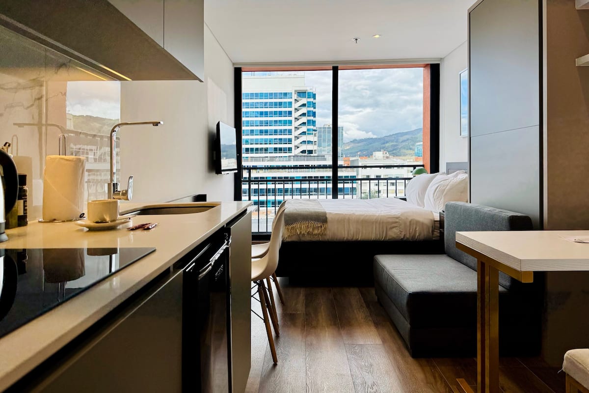 A modern hotel room with a kitchen area, a bed with a view of a cityscape, and a balcony.