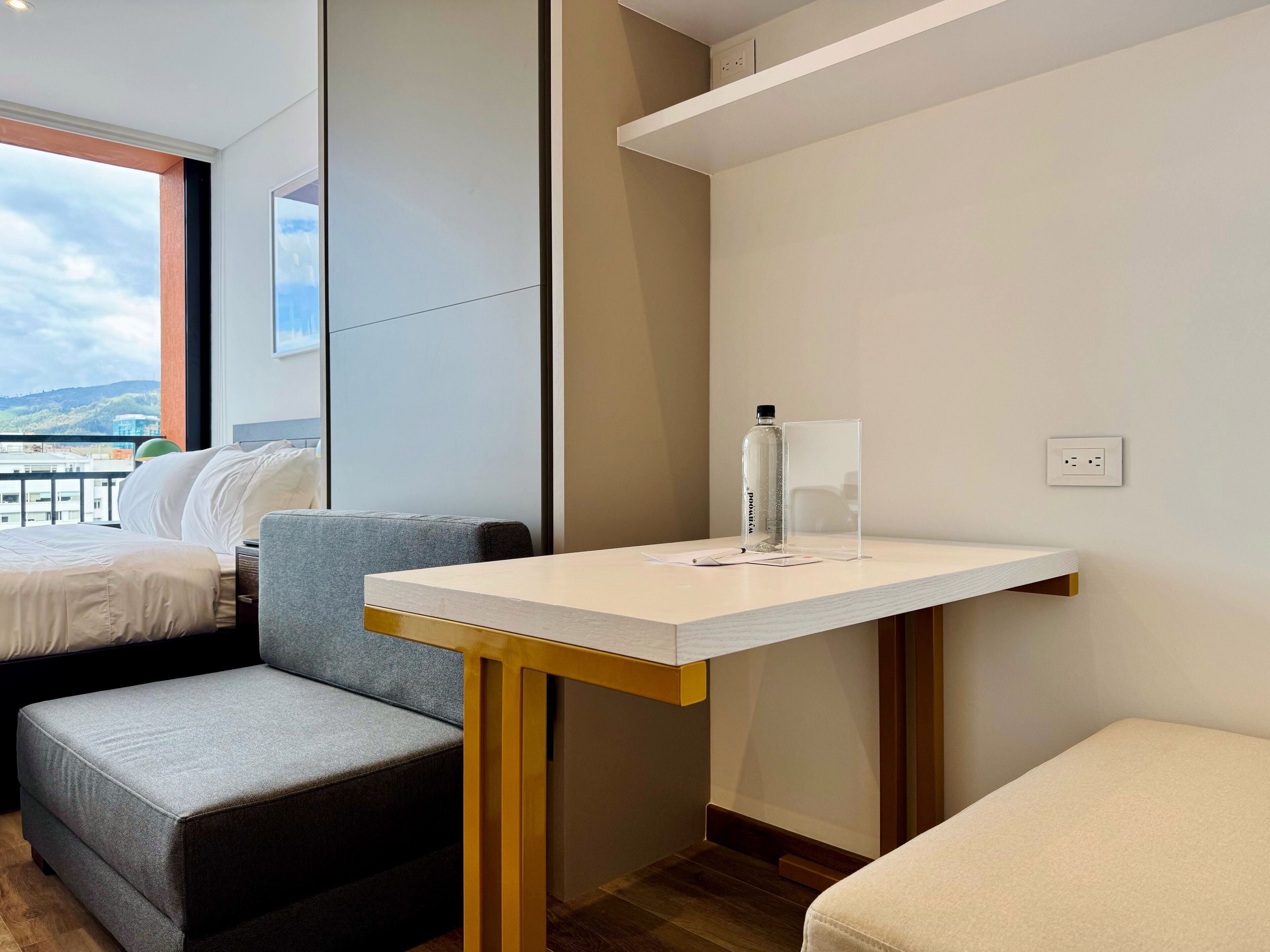 A modern hotel room with a bed, a small sofa, a table with a bottle and a glass, and a view of the outdoors.