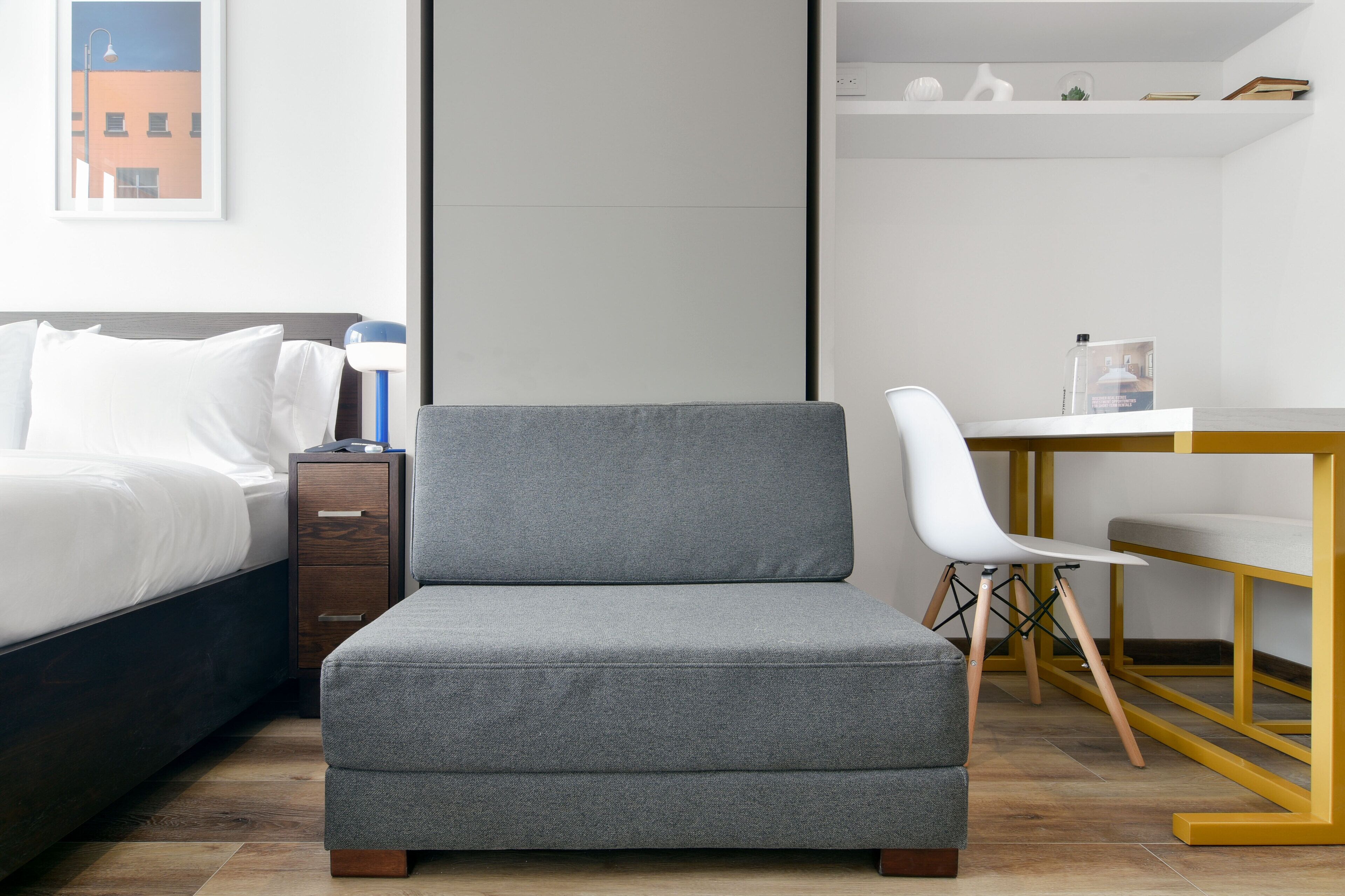 A modern bedroom with a bed, a grey armchair, a white chair, a small table, and a bookshelf.