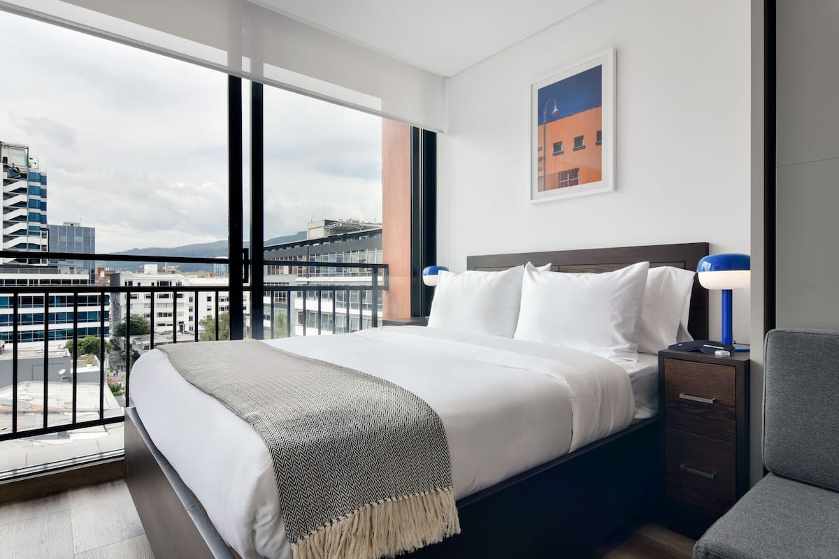 A hotel room with a large window, a bed with white linens, a nightstand with a lamp, and a view of the cityscape.