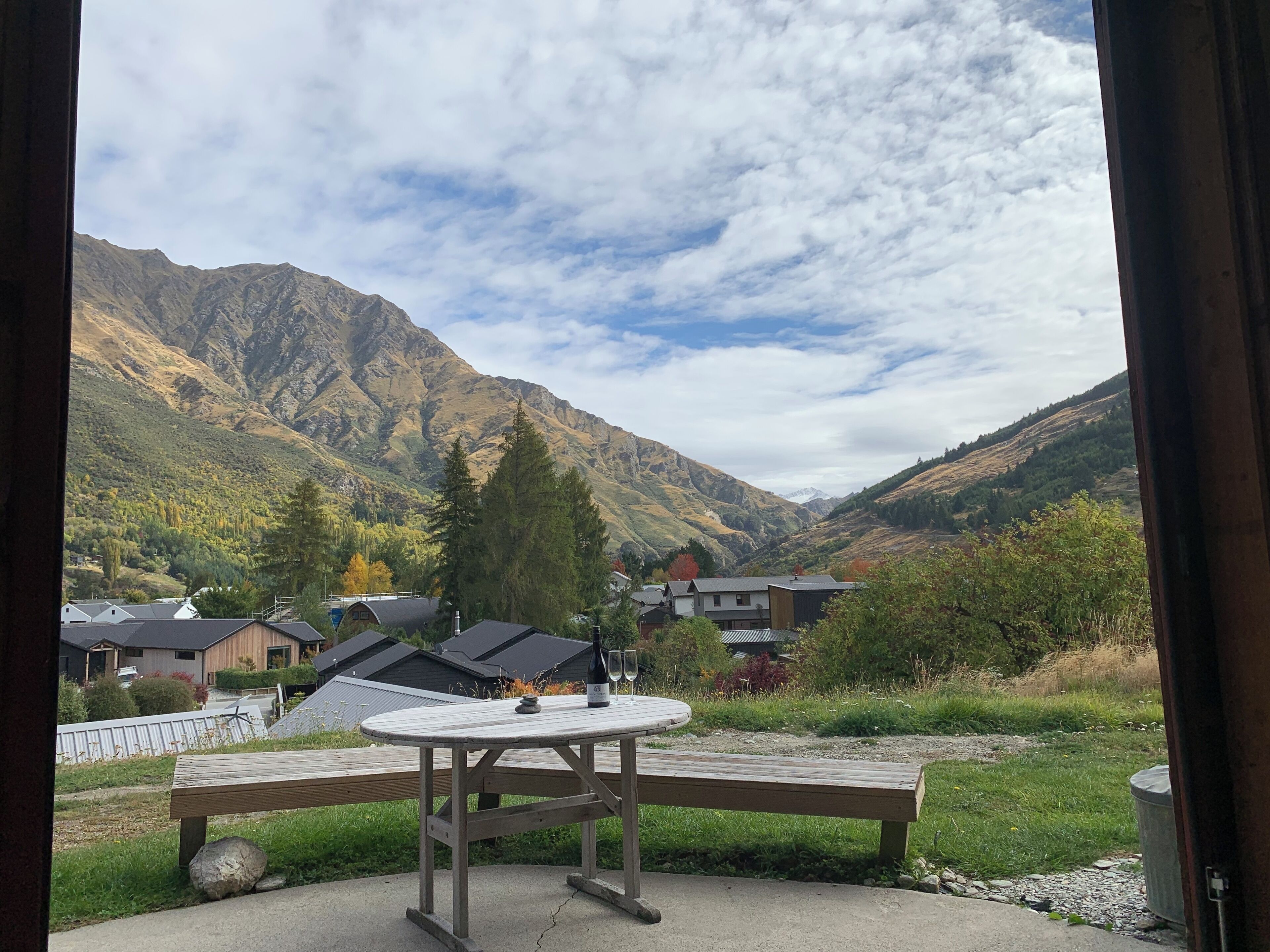 A view from inside a building looking out at a mountainous landscape with houses and a picnic table with a bottle and glass.