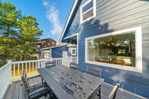 A deck with a table and chairs, a large window, and a blue house. - NEW! Seaside Castle | Beach | Promenade | Hot tub (Seaside)