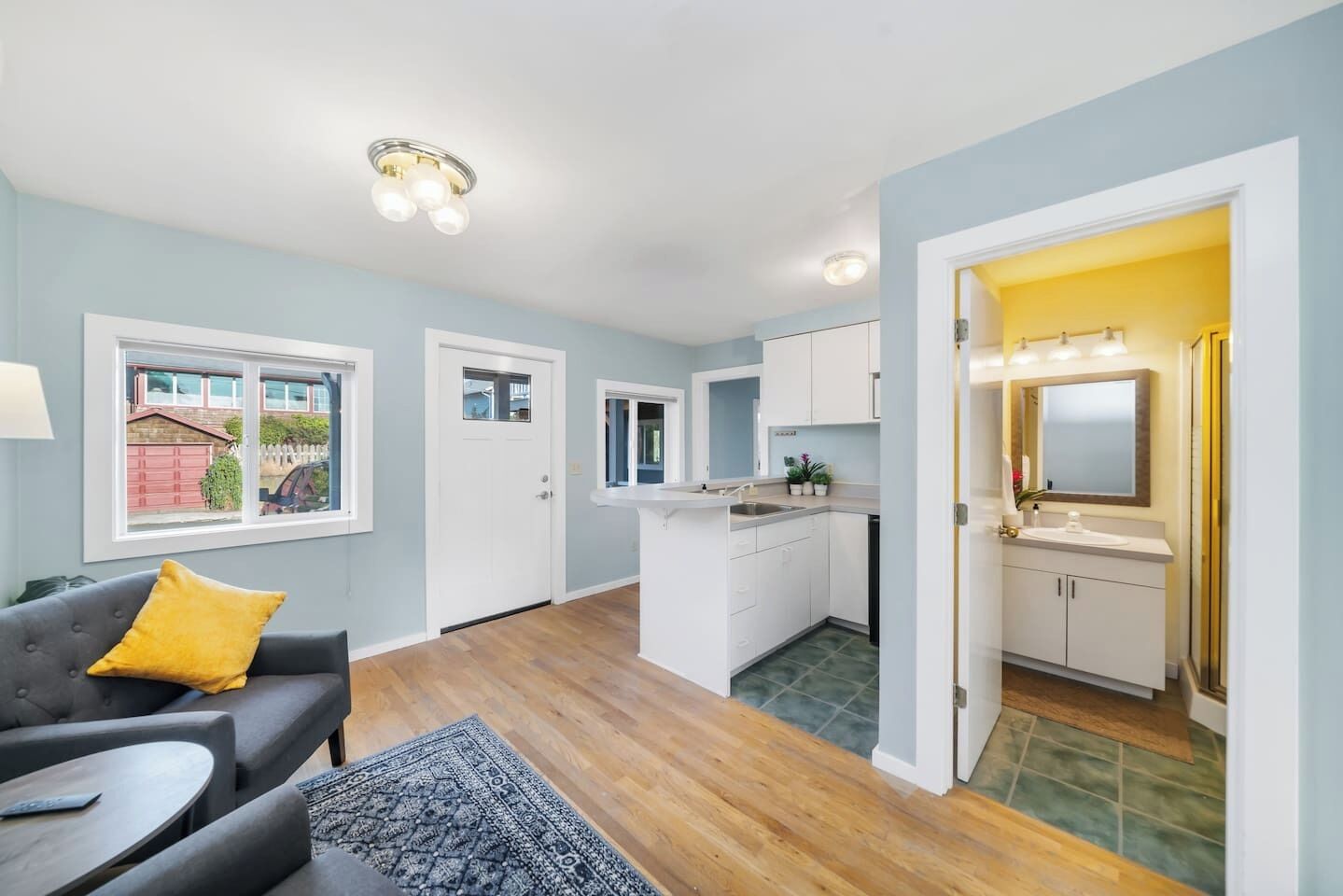 Eine moderne Wohnung mit Wohnzimmer, Küche und Bad. Es gibt ein Sofa mit einem gelben Kissen, einen Esstisch und eine Kücheninsel. Das Apartment bietet Blick auf eine rote Garage und ein Haus.
