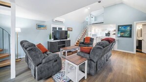 A modern living room with grey sofas, a coffee table, and a TV. There is a staircase leading to an upper level.