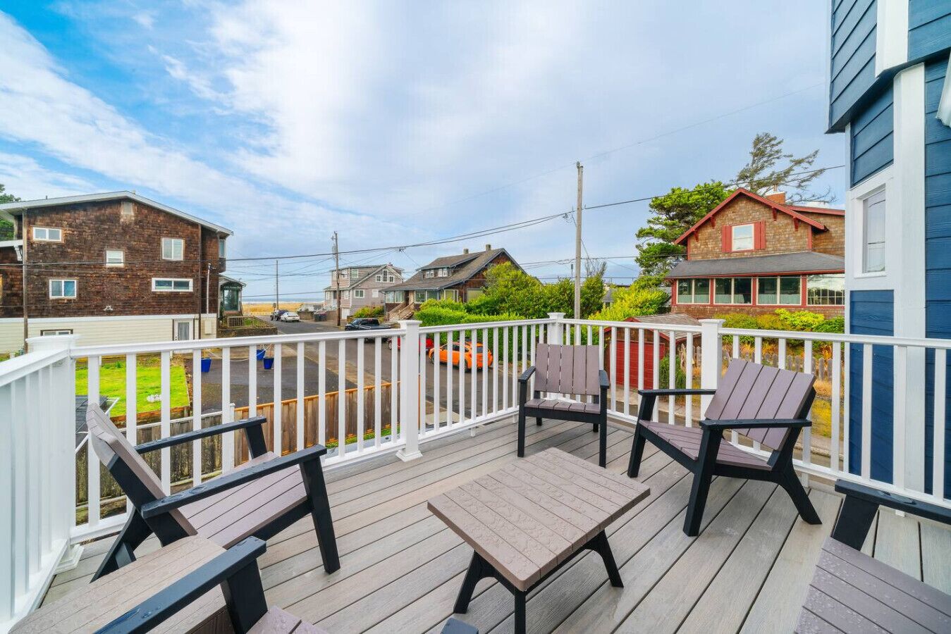 A wooden deck with two chairs and a table, surrounded by a white railing, overlooking a residential street.