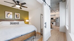 A modern hotel room with a bed, ceiling fan, artwork, and a view into the kitchen area. - Beltline Lux Loft (Atlanta)